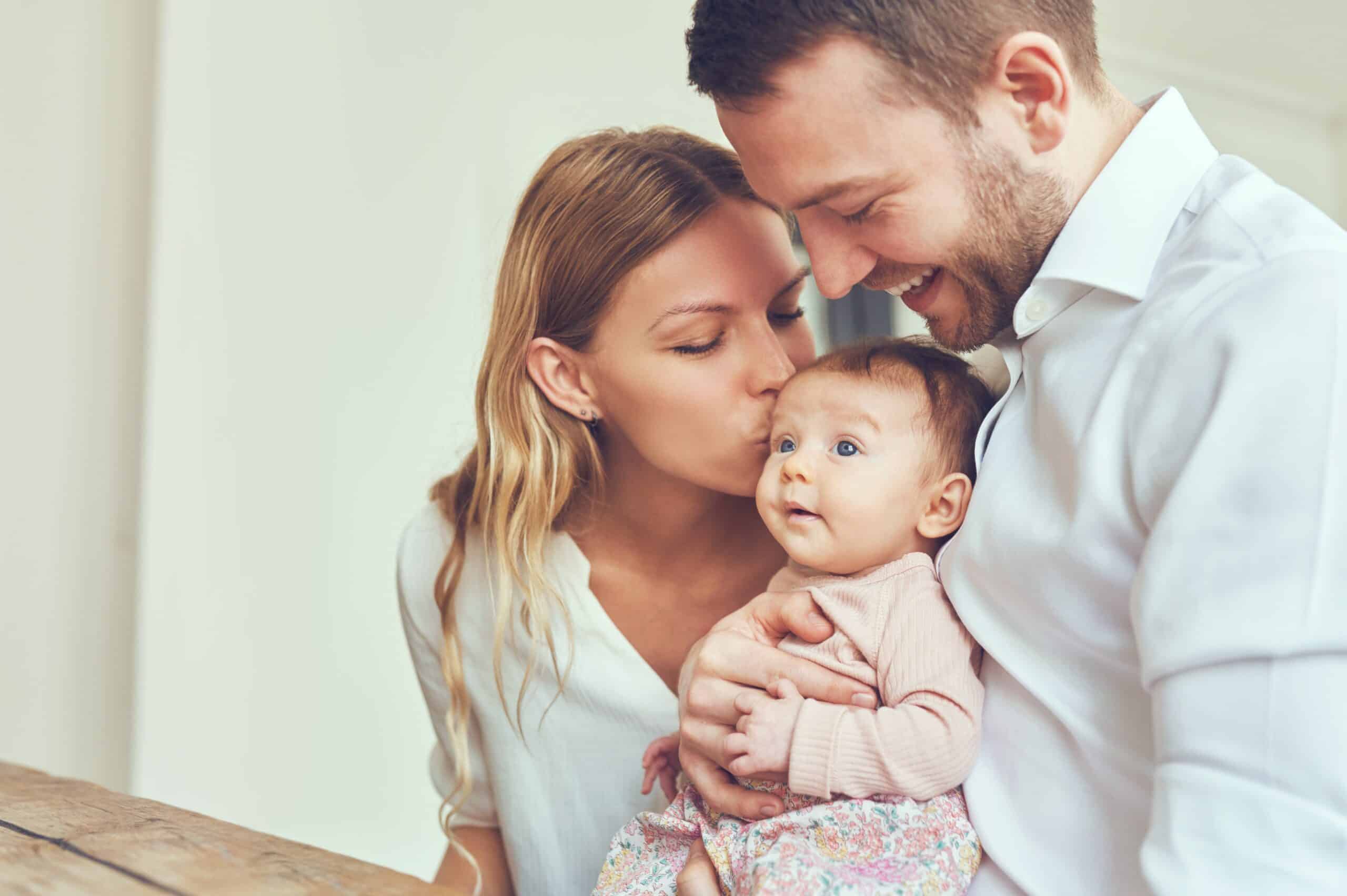 A mother and father smile warmly as they hold their baby; the mother gently kisses the infant's forehead in a bright, cozy indoor setting.